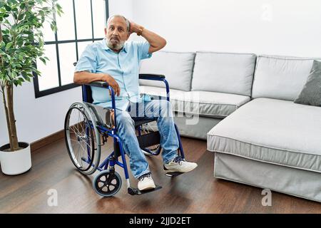 Hübscher älterer Mann, der im Wohnzimmer auf dem Rollstuhl sitzt, verwirrt und wundert sich über Fragen. Unsicher mit Zweifel, mit der Hand auf dem Kopf denkend. Stifte Stockfoto