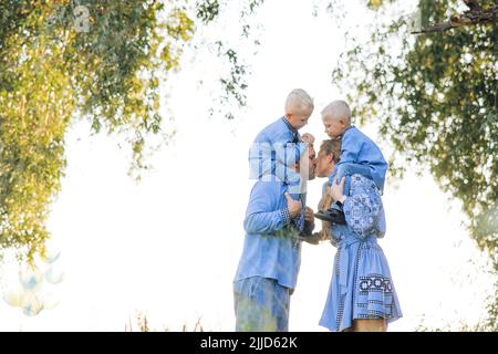 Glückliche Eltern tragen ihre beiden Zwillingsöhne auf ihren Schultern und küssen sich beim Spaziergang auf der Wiese. Sie sind in ukrainischem nationalem Gestrick gekleidet Stockfoto