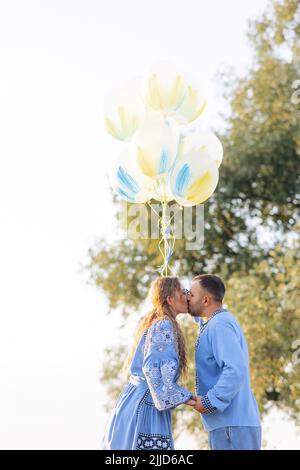 Glückliches Paar küsst sich gegenseitig und hält Luftballons während des Spaziergangs auf der Wiese. Sie sind in ukrainische nationale bestickten Hemden gekleidet. Stockfoto