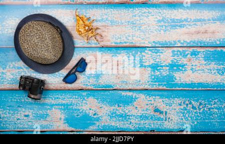 Urlaubszubehör auf den blauen Holzbrettern. Lage: Rechte Ecke. Reisehut, Kamera, Sonnenbrille, große Muschel. Stockfoto