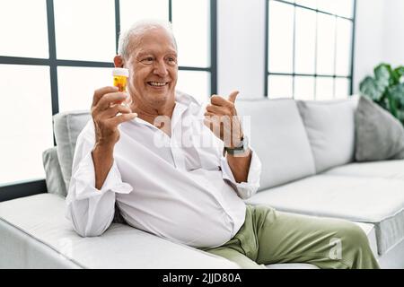 Älterer Mann hält Pillen lächelnd mit glücklichem Gesicht und zeigt mit dem Daumen nach oben zur Seite. Stockfoto