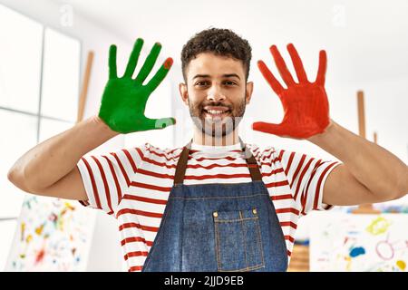 Junger arabischer Künstler lächelt glücklich und zeigt gemalte Hände im Kunststudio. Stockfoto