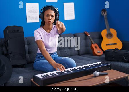 afroamerikanische Frau mit Zöpfen, die Klaviertastatur im Musikstudio spielen, zeigt mit dem Finger auf die Kamera und auf Sie, selbstbewusst Geste aussehende ser Stockfoto