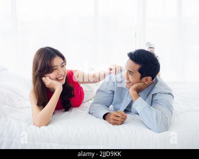 Der süße Moment des glücklichen Paares, das auf dem weißen Bett liegt. Asiatischer Mann im Denim-Hemd und junge schöne Frau lächeln und unterhalten sich auf dem Bett in Weiß Stockfoto