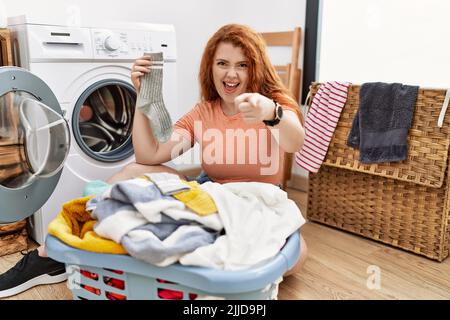 Junge Rothaarige Frau, die schmutzige Wäsche in die Waschmaschine legt und mit glücklichen und lustigen Gesichtes die Finger zur Kamera zeigt. Gute Energie und Vibes. Stockfoto