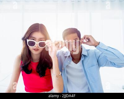 Glückliches asiatisches Paar im Sommer, junger Mann und Frau tragen eine Sonnenbrille, berühren sie und schauen auf Kamera auf dem Bett im hellen Schlafzimmer in der Nähe des Glases Stockfoto