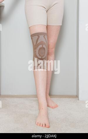 Kniestützenstrebe auf einem Frauenbein. Mädchen in einer Orthese im Inneren des Hauses. Orthopädische anatomische Hosenträger zur Kniefixierung, Verletzungen und Schmerzen Stockfoto