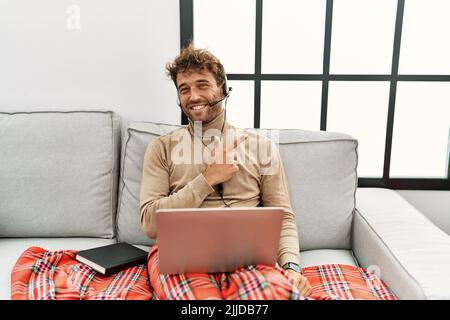Ein junger, gutaussehender Mann mit Bart, der ein Headset trägt und von zu Hause aus fröhlich mit einem Lächeln im Gesicht arbeitet, das mit Hand und Finger zur Seite zeigt Stockfoto