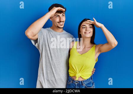 Junge hispanische Paar, die zusammen über blauem Hintergrund stehen, sehr glücklich und lächelnd und weit weg mit der Hand über dem Kopf schauen. Suchkonzept. Stockfoto