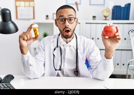 afroamerikanischer Arzt hält verschreibungspflichtige Pillen und frischen Apfel im Schockgesicht, sieht skeptisch und sarkastisch aus, überrascht mit offenem Mund Stockfoto
