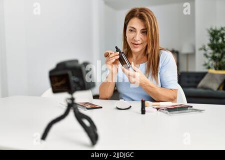 Kaukasische Frau mittleren Alters macht Make-up-Tutorial mit Kamera sitzen auf dem Tisch zu Hause. Stockfoto