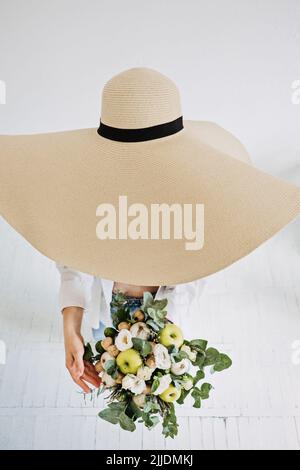 Draufsicht auf Frau in großen Strohhut mit Blumenstrauß in der Hand. Gesichtslose Studioaufnahme einer Frau Stockfoto