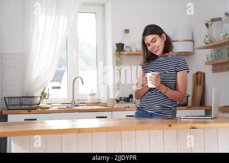 Eine Frau in der Küche mit einem Lächeln. Eine Frau trinkt Kaffee und hält eine Tasse in der Hand. Sie freut sich, weil sie morgens ihren Lieblingskaffee trinkt. Hochwertige Fotos mit Platz für Kopien. Stockfoto