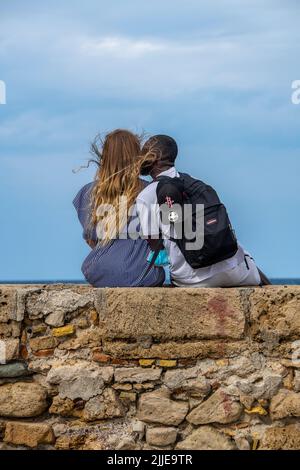 Junge Mischrenner, die an einer Wand am Meer sitzen, Mischrenner, die an einer Wand sitzen, Paare, die sich am Strand, am Uferdamm, an der Promenade umarmen, Stockfoto
