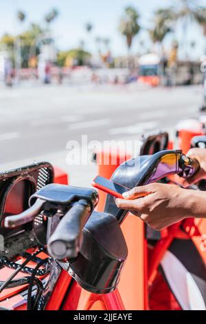 Nahaufnahme der Hände einer nicht erkennbaren afrikanischen Frau. Sie benutzt ihr Telefon, um das öffentliche System des Fahrradverleihens zu nutzen Stockfoto