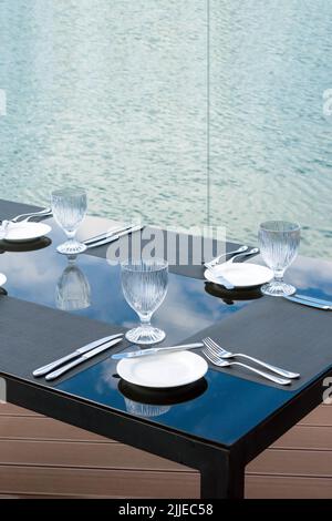 Leere Gläser, weiße Teller und Besteck auf einem Tisch in einem Strandrestaurant. Tischstimmung für ein festliches Abendessen. Stockfoto
