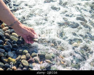 Eine Hand mit einem Armband aus Steinen hält einen Kieselstein in der Nähe des Wassers. Meereswelle und weibliche Hand. Spüren Sie die Welle. Kieselstrand. Seeufer Stockfoto