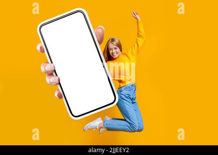 Fröhliche Dame springt in der Luft auf orangenen Hintergrund, zeigt Smartphone Stockfoto