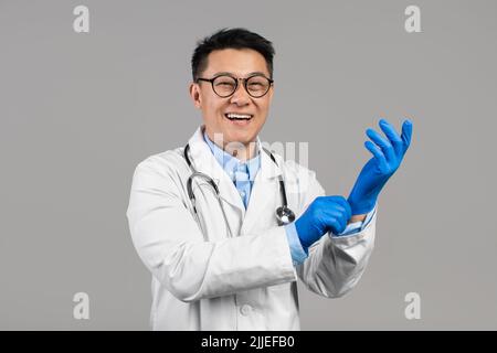 Fröhlicher, tausendjähriger chinesischer Arzt im weißen Mantel, Brille mit Stethoskop trägt Gummihandschuhe Stockfoto