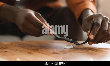 Junger afroamerikanischer Kerl, der eine moderne Smartwatch zum Aufladen auf einem kabellosen Ladegerät auf einem Holztisch ablegt, Nahaufnahme Stockfoto