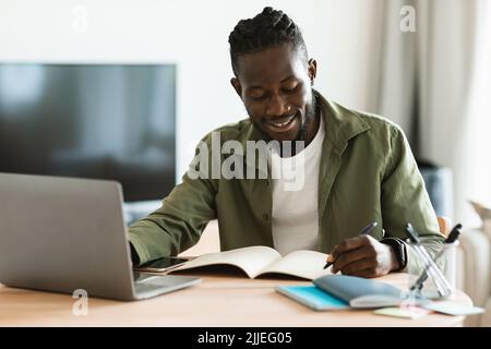 Lächelnder afroamerikanischer Mann, der am Schreibtisch sitzt, am Laptop arbeitet und sich im Notizbuch Notizen macht, schwarzer Mann, der online studiert Stockfoto