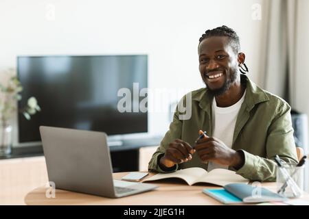 Home Office-Konzept. Glücklicher afroamerikanischer Mann, der am Schreibtisch sitzt, an einem Laptop arbeitet und Notizen schreibt, lächelt der Kamera zu Stockfoto