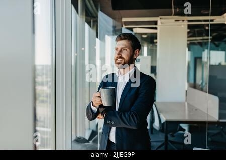 Ein gutaussehender, erfolgreicher Geschäftsmann, der eine Kaffeepause macht und neben dem Fenster in seinem Büro mit einer Tasse Kaffee steht Stockfoto