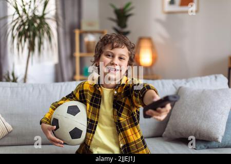 Kleiner Fußballfan, der zu Hause fernsehen schaut, auf dem Sofa mit Fernbedienung und Fußballball sitzt und vor der Kamera lächelt Stockfoto