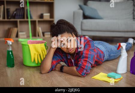 Traurige müde tausendjährige afroamerikanische Frau Hausfrau mit Reinigungsmitteln auf dem Boden liegen Stockfoto
