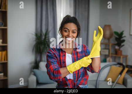 Lächelnde junge afroamerikanische Frau zieht Gummihandschuhe an und ist bereit, im Wohnzimmer Hausaufgaben zu machen Stockfoto