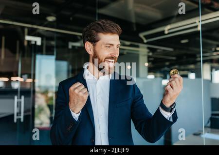 Ein gutaussehender, glücklicher Geschäftsmann, der Bitcoin hält und stolz schreit und den Sieg und den Erfolg im Amt feiert Stockfoto