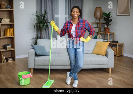 Glückliche junge afrikanisch amerikanische Frau Hausfrau mit Mop Reinigung Boden in gemütlichen Wohnzimmer Interieur Stockfoto