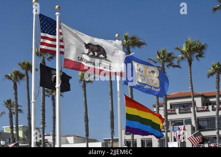 Mehrere Flaggen fliegen am Huntington Beach vor Palmen vor einem wolkenlosen blauen Himmel, darunter Stars & Stripes und kalifornische Flaggen. Stockfoto