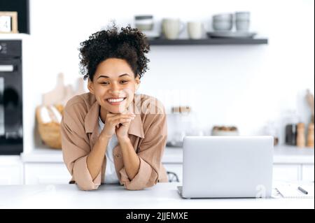 Schöne positive selbstbewusste afroamerikanische lockige junge Frau, in einem beigen Casual Shirt, steht in der Küche am Tisch neben dem Laptop, schaut auf die Kamera, lächelt glücklich Stockfoto