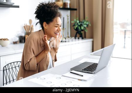 Aufgeregte, fröhliche afroamerikanische junge Frau, die zu Hause in der Küche sitzt, ihren Laptop benutzt, erstaunt auf den Bildschirm schaut, mit den Armen eine Siegergeste macht, den Erfolg feiert und glücklich lächelt Stockfoto