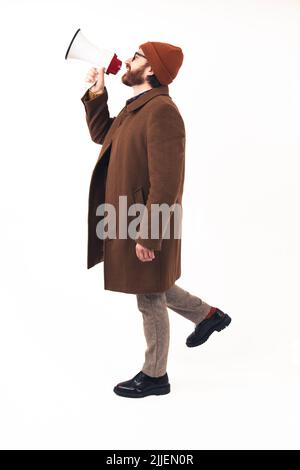 Ein Mann in seinem Alter von 30s Jahren, der mit einem Megaphon schrie - gekleidet in einen Kamelmantel und stilvolle Stiefel - Profil auf weißem Hintergrund aufgenommen - isoliert. Hochwertige Fotos Stockfoto