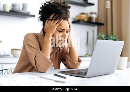 Verzweifelt besorgt traurig afroamerikanische junge Frau, mit Laptop für die Arbeit oder Studium von zu Hause aus, sitzen in der Küche, frustriert Blick auf den Bildschirm, gescheitert Projekt, bekam finanziellen Verlust, Arbeitsplatz Downgrade Stockfoto