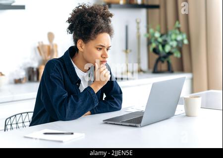 Eine ängstliche afroamerikanische Frau, Kauffrau, Freiberuflerin, schaut besorgt auf den Laptop-Bildschirm, beobachtet die Situation auf dem Finanzmarkt, analysiert konzentriert die Dynamik, fühlt sich stressig Stockfoto