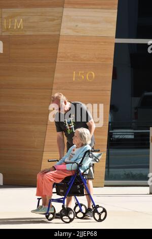 Ein kaukasischer älterer Mann, der einer älteren Frau hilft, die an einem sonnigen Tag auf dem Rollator-Rollstuhl vor dem Gebäude sitzt. Stockfoto