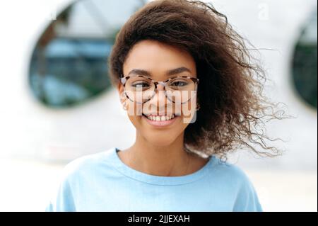 Nahaufnahme einer selbstbewussten, erfolgreichen, jungen, lockigen afroamerikanischen Geschäftsfrau oder Schülerin, die mit einer Brille im Freien steht und mit einem süßen Lächeln auf die Kamera blickt Stockfoto