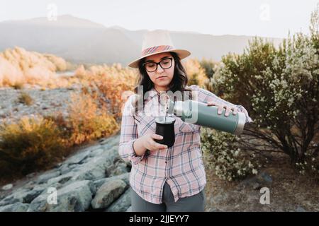 Junge latinerin mit Hut, serviert Mate mit einem hellgrünen Thermo, in einem natürlichen Raum. Stockfoto