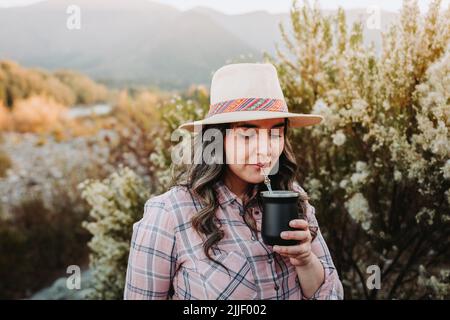 Eine Latinerin mit Hut und einer blassrosa Bluse, trinkende Partnerin in einem natürlichen Raum bei Sonnenuntergang Stockfoto