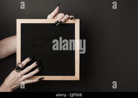 Halloween-Nachbau. Briefbrett in Händen mit langen schwarzen Nägeln und Spinnenring auf schwarzem Hintergrund. Vorderansicht. Stockfoto