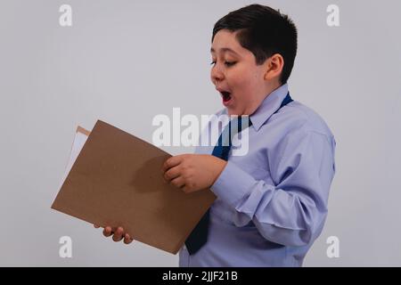 Ein Junge in einem eleganten Hemd und Krawatte, der vorgibt, ein Geschäftsmann zu sein, während er mit einem überraschten Gesicht einige Papiere liest. Der Junge, der als Erwachsener mit Hemd und Krawatte verkleidet ist, liest einen Vertrag in einem Ordner. Stockfoto