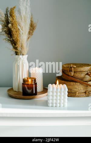 Inneneinrichtung mit Blumenmuster. Getrocknete Blumen in Vase auf weißem Regal und Holztablett mit verschiedenen hausgemachten Kerzen Stockfoto