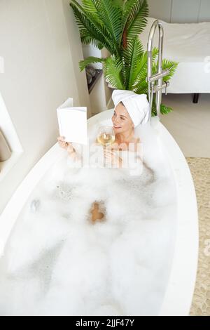 Attraktive kaukasische junge Frau mit Handtuch auf dem Kopf, Baden und Buch mit Wein lesen. Stockfoto