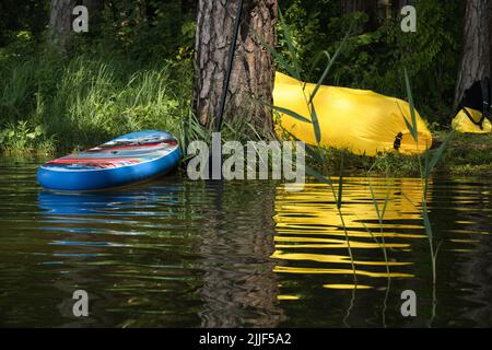 Stand up Paddleboard und gelbe aufblasbare Liege am Ufer des Sees im Schatten der Bäume und Reflexionen im ruhigen Wasser Stockfoto