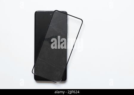 Telefon und gebrochenes Schutzglas liegen in der Nähe auf einem weißen Hintergrund, Austausch von Schutzglas auf einem Smartphone Stockfoto