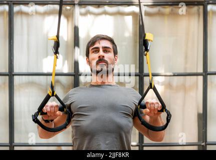 Entschlossener bärtiger Sportler im Aktivbekleidung, der Liegestütze mit TRX-Seilen in der Nähe von Fenstern während des intensiven Workouts in einem modernen, hellen Fitnessstudio macht Stockfoto