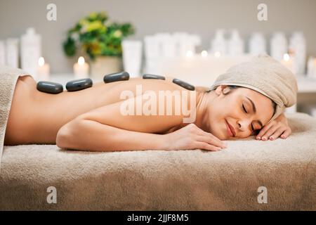 Eine sehr erfreuende Kundin mit heißen Steinen auf dem Rücken, die während der Massage im modernen, hellen Spa auf der Couch mit Handtuch lag Stockfoto